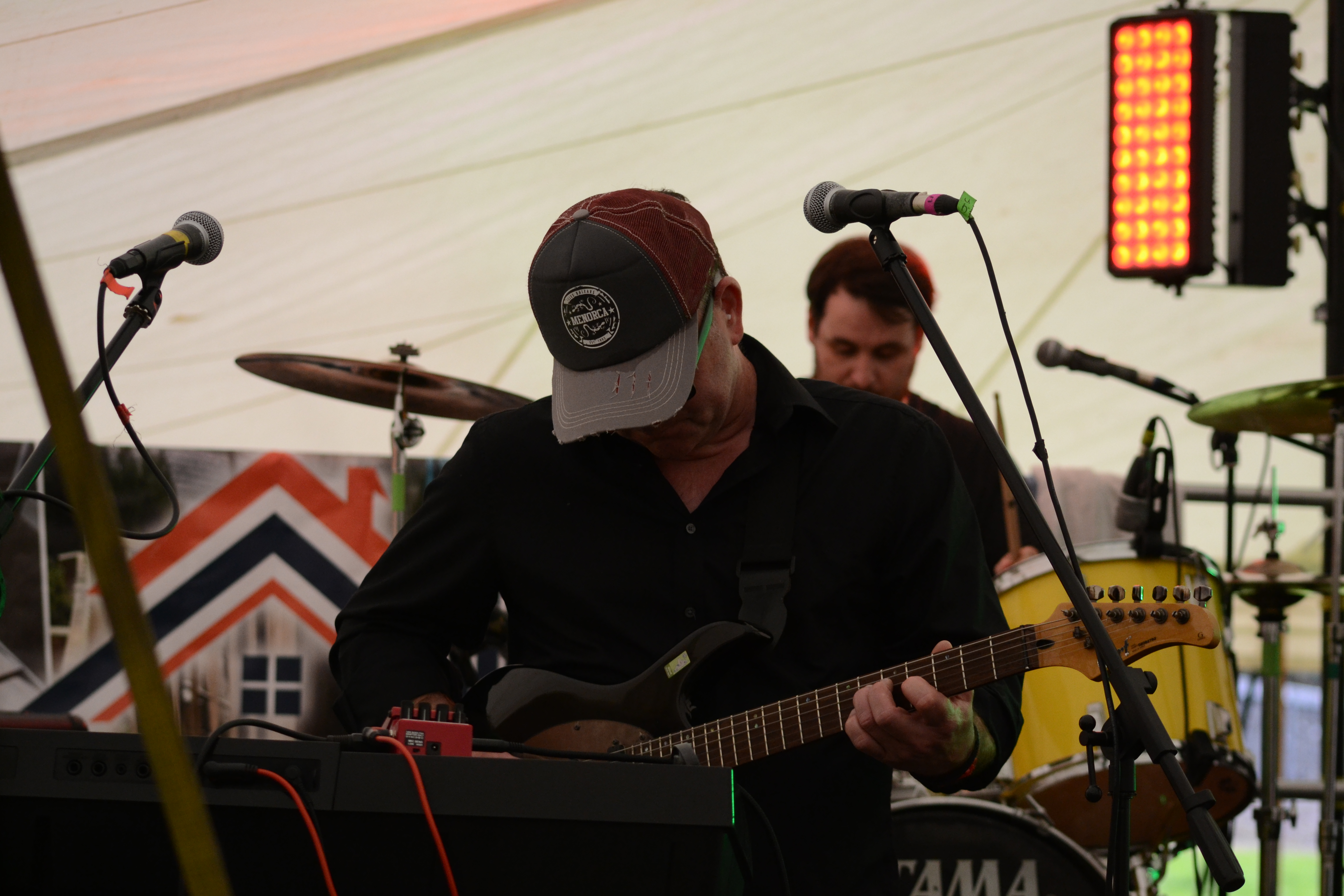 Guitarist in cap and sunglasses singing while playing black electric guitar, drummer behind