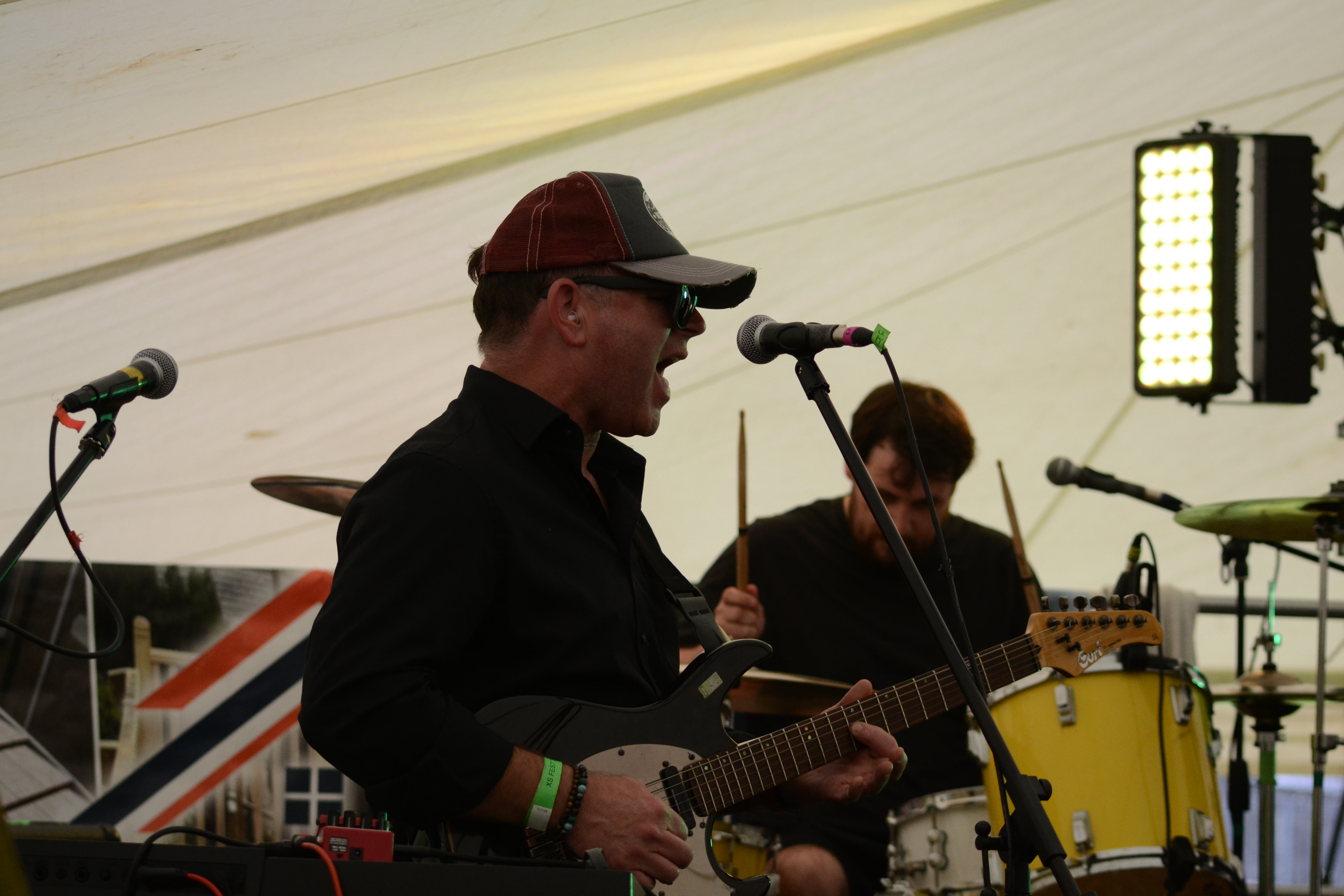 Wide full-band shot with guitarist left, drummer center, and bassist adjusting at right