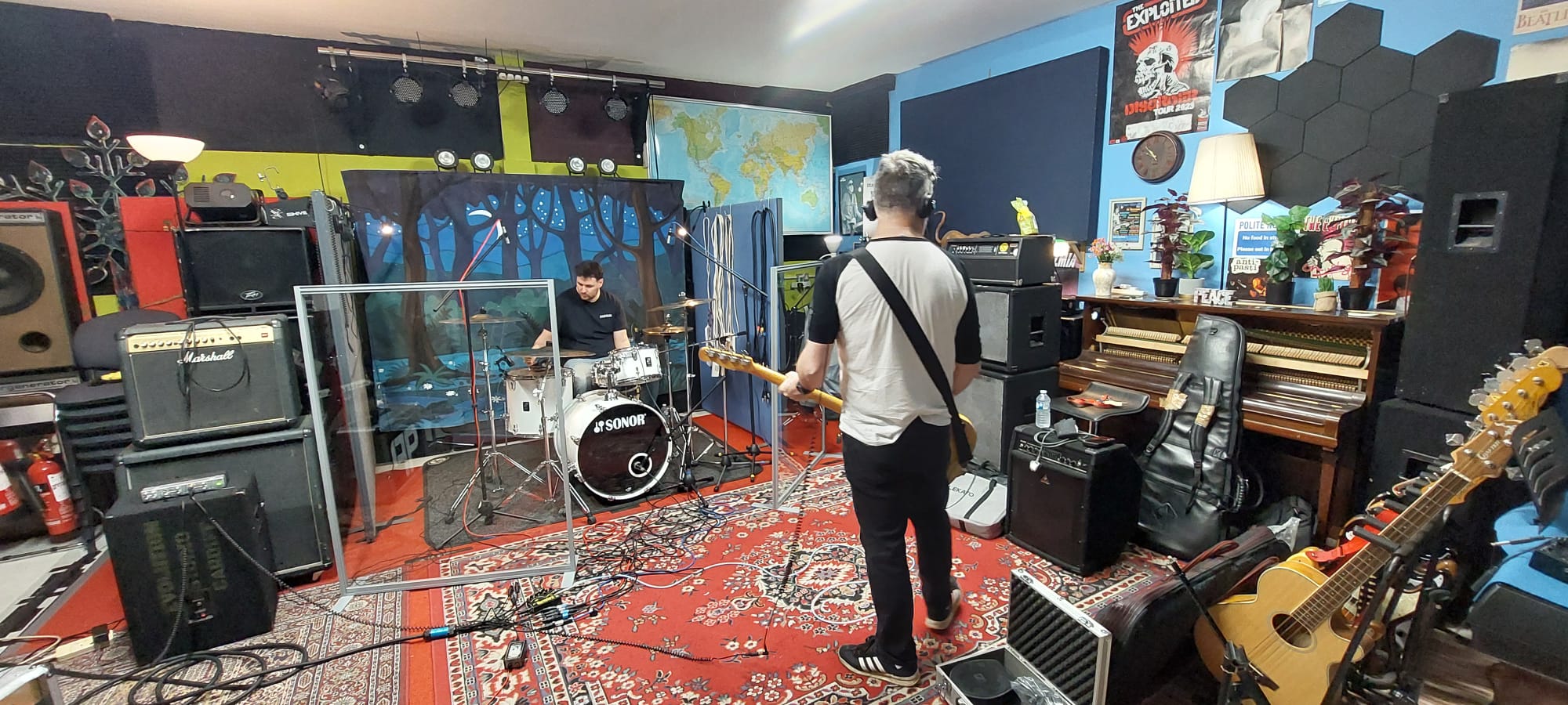 Wide studio view with bassist facing drummer behind acoustic screens