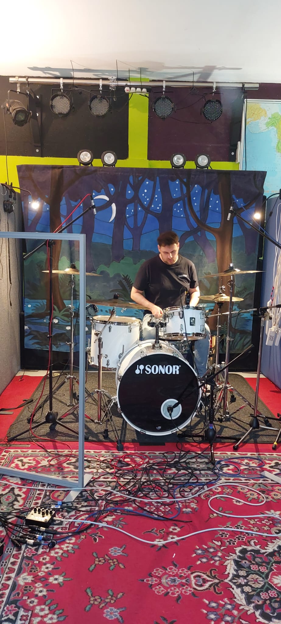 Drummer playing a white Sonor kit with overhead mics and isolation screen