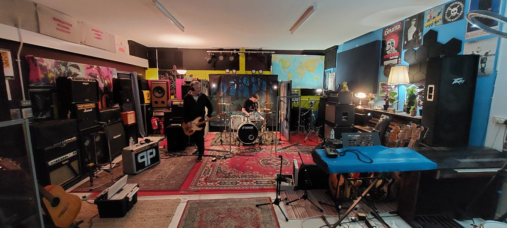 Wide room shot of bassist and drummer recording among amps and mic stands